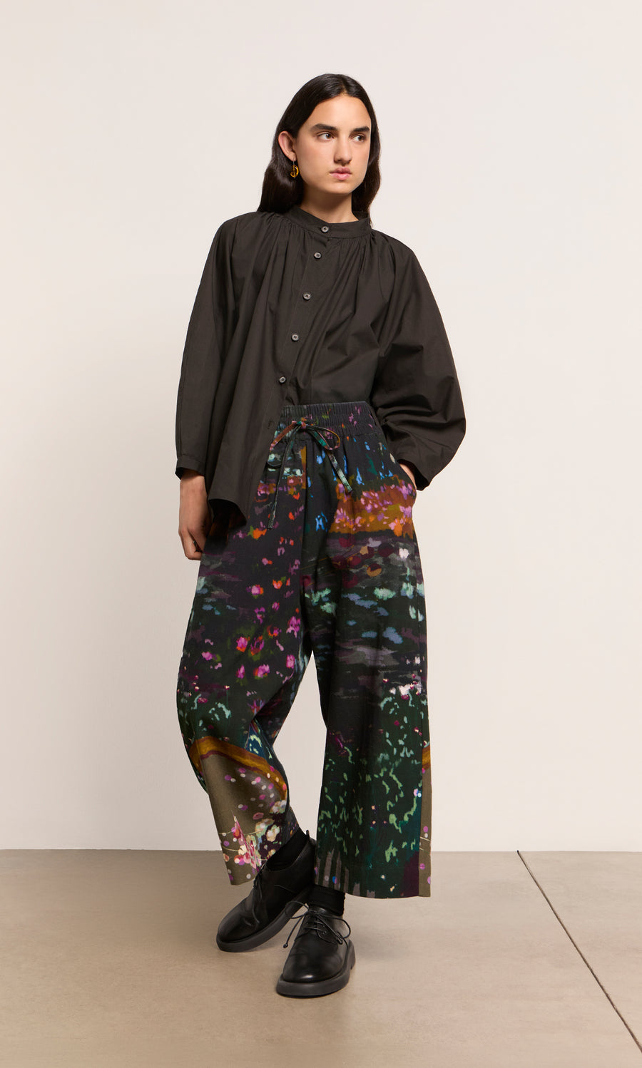 Plümo  Person in a Kroyer Blouse with balloon sleeves and vibrant floral wide-leg pants stands with one foot forward.