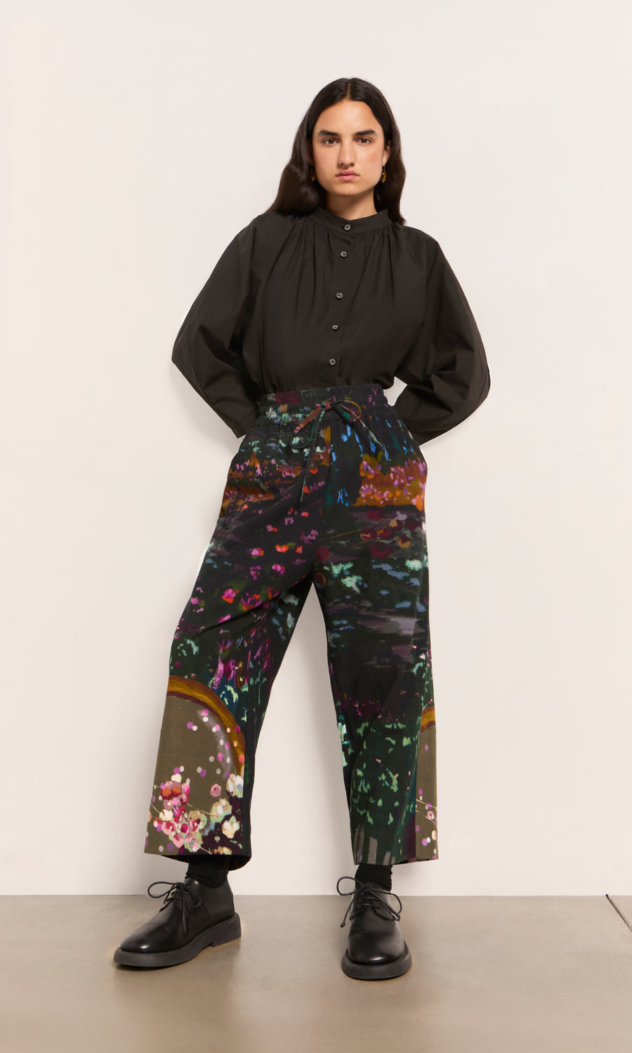 Plümo  Person in a dark brown Kroyer Blouse and colorful wide-leg pants stands by a plain wall, hands tucked in pockets.