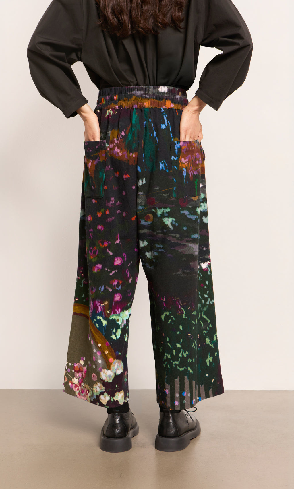 Plümo  Person in Water Colour Pants with jewel-toned print, black shoes, hands on hips, seen from the back.