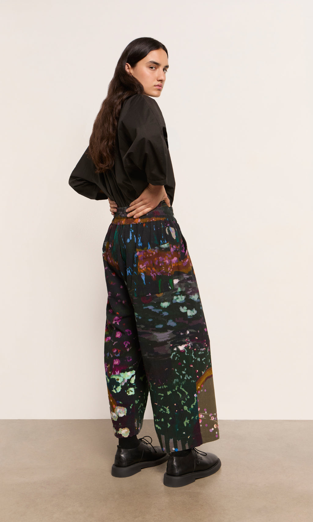 Plümo  Person with long hair wears a black shirt and Water Colour Pants, standing sideways and looking back.