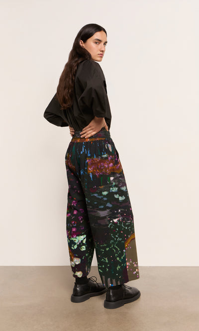 Plümo  Person with long hair wears a black shirt and Water Colour Pants, standing sideways and looking back.