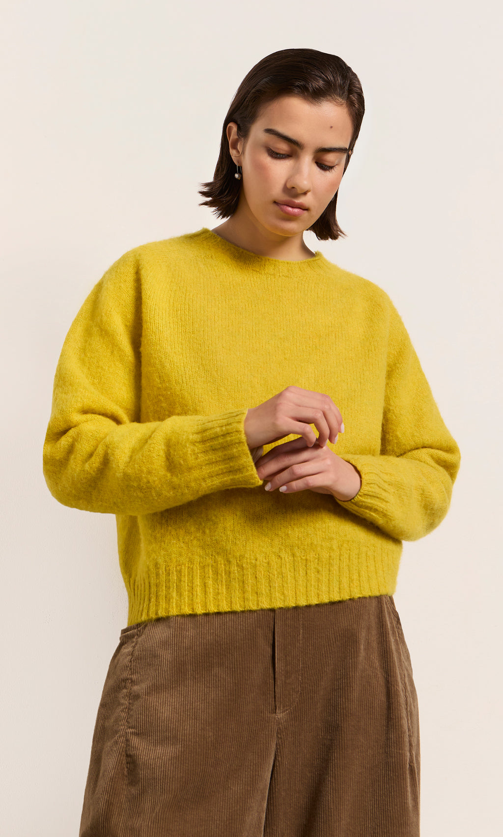 Plümo  Person in a Wicklow Crewneck - Golden Hop sweater and brown pants, looking down with hands together.