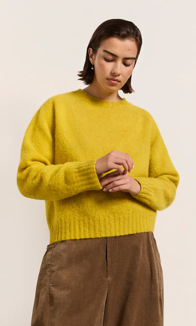 Plümo  Person in a Wicklow Crewneck - Golden Hop sweater and brown pants, looking down with hands together.