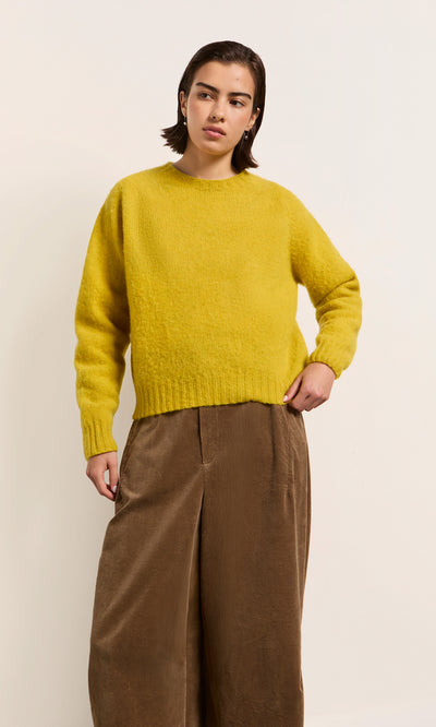 Plümo  Model in the Wicklow Crewneck - Golden Hop stands, gazing left, wearing wide brown pants against a plain backdrop.