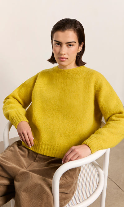 Plümo  Person with short dark hair wears the Wicklow Crewneck - Golden Hop, sitting on a white chair in tan corduroy pants.