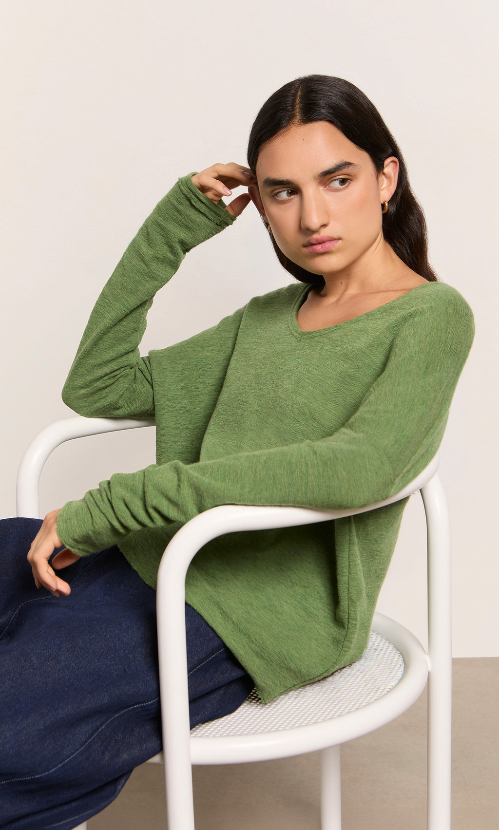 Plümo  Person with long dark hair sits on a white chair, wearing the marled green Wisher Jumper and dark pants, side-facing.