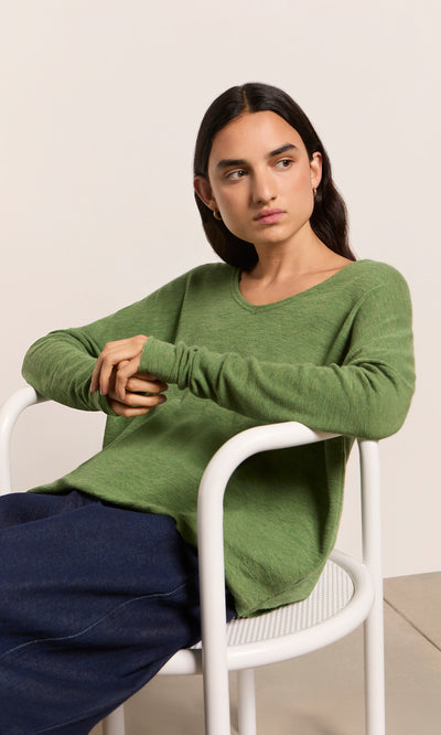 Plümo  Person seated on a white chair in the Wisher Jumper, marled green, dark pants—showcasing Danish design elegance.