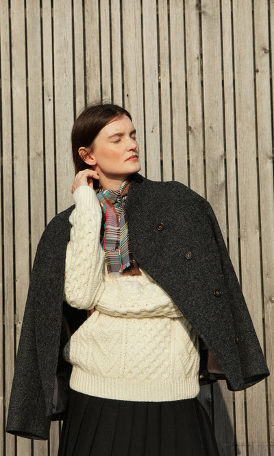 Plümo- Woman in a Berg Aran Sweater and scarf, with a dark coat over her shoulders, stands by a wooden fence.