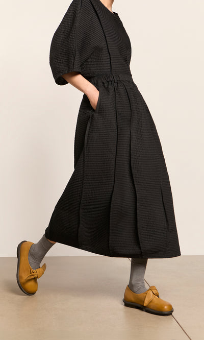 Plümo  Person in a black Yoshiko Bubble Skirt, gray socks, mustard yellow shoes, walking indoors.
