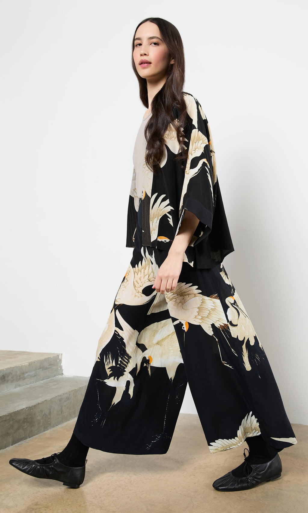 Plümo- Woman wearing a Black Stork Kimono with relaxed fit walks indoors by stairs, her long hair loose.