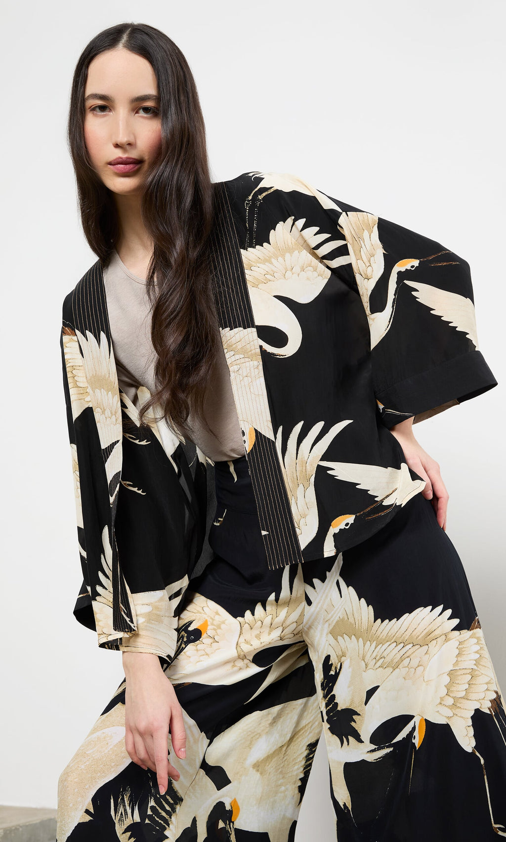 Plümo- Woman wears Black Stork Kimono, posing with one hand on her knee against a plain background.