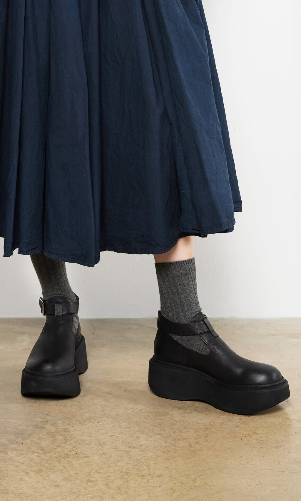 Plümo- Person wearing a navy skirt, Lambswool Socks - Graphite, and black platform Mary Janes on a concrete floor.