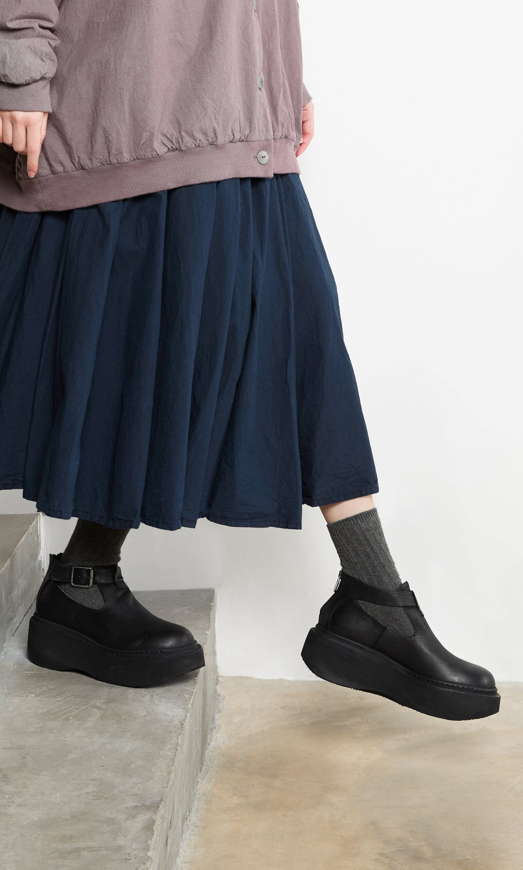 Plümo- Person in a navy skirt, Lambswool Socks - Graphite, and black platform shoes steps down concrete stairs.