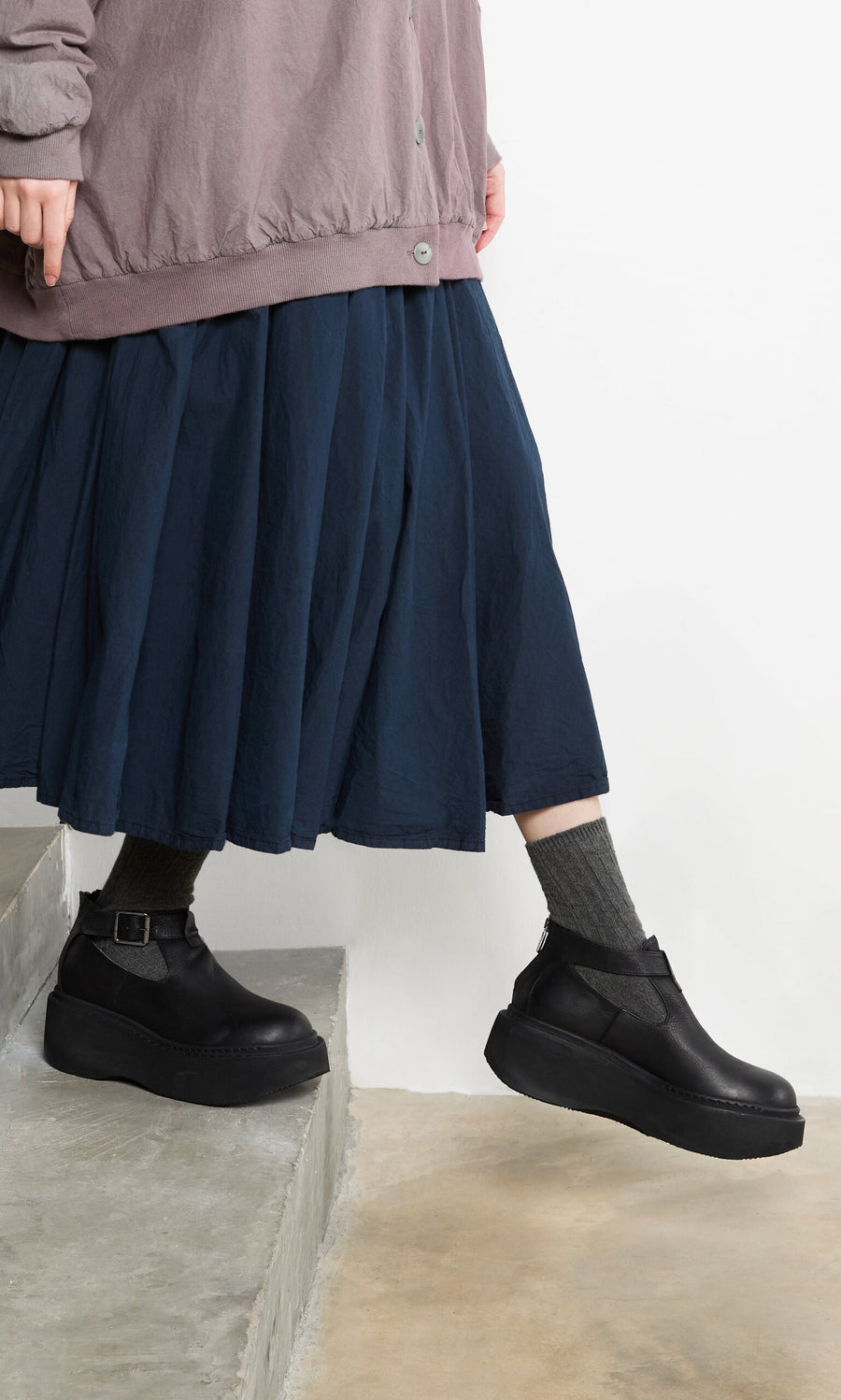 Plümo- Person in a navy skirt, Lambswool Socks - Graphite, and black platform shoes steps down concrete stairs.