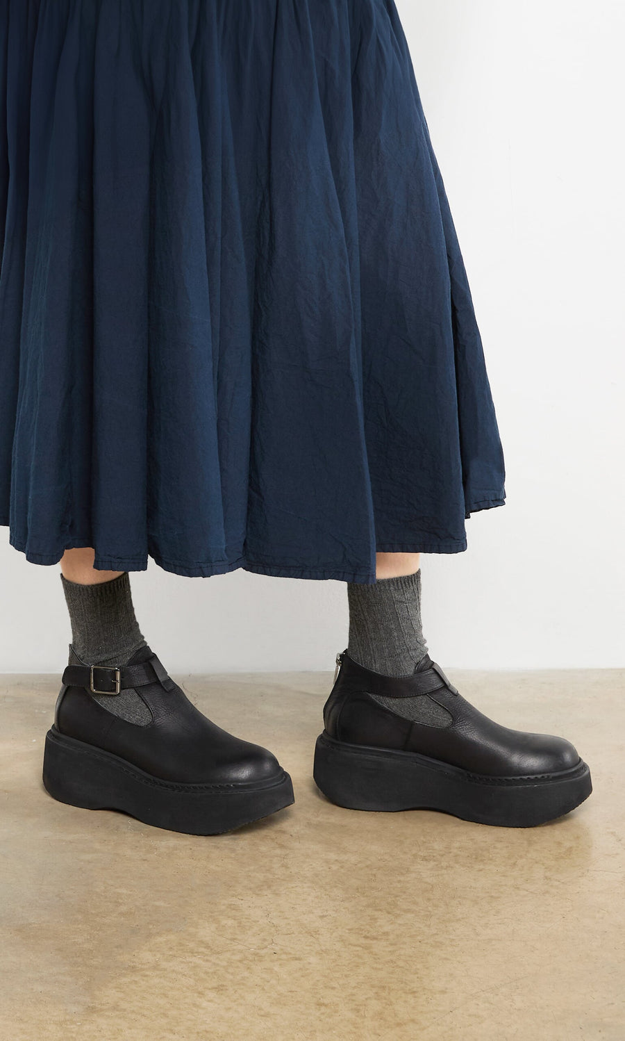 Plümo- Person wears a navy skirt, Lambswool Socks - Graphite, and black platform strap shoes on a beige floor.