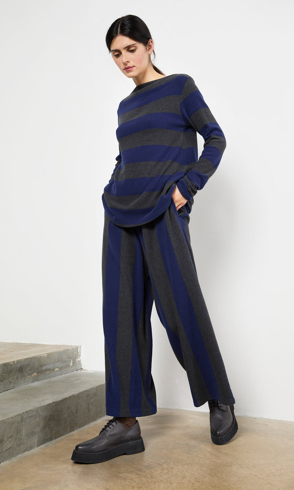 Plümo- Woman in Clara Stripe Pants stands near concrete steps, pairing them with a loose blue and gray striped sweater.