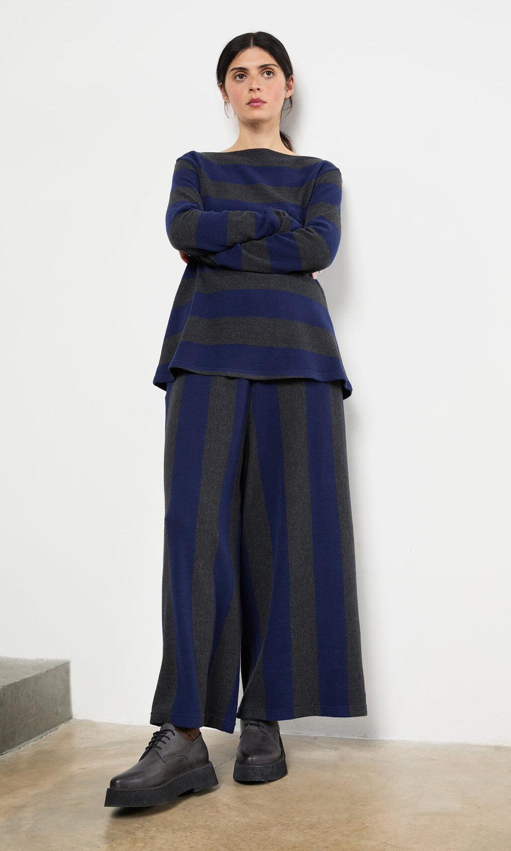 Plümo- Woman standing against a white wall wearing Clara Stripe Pants and a loose striped top with black shoes.