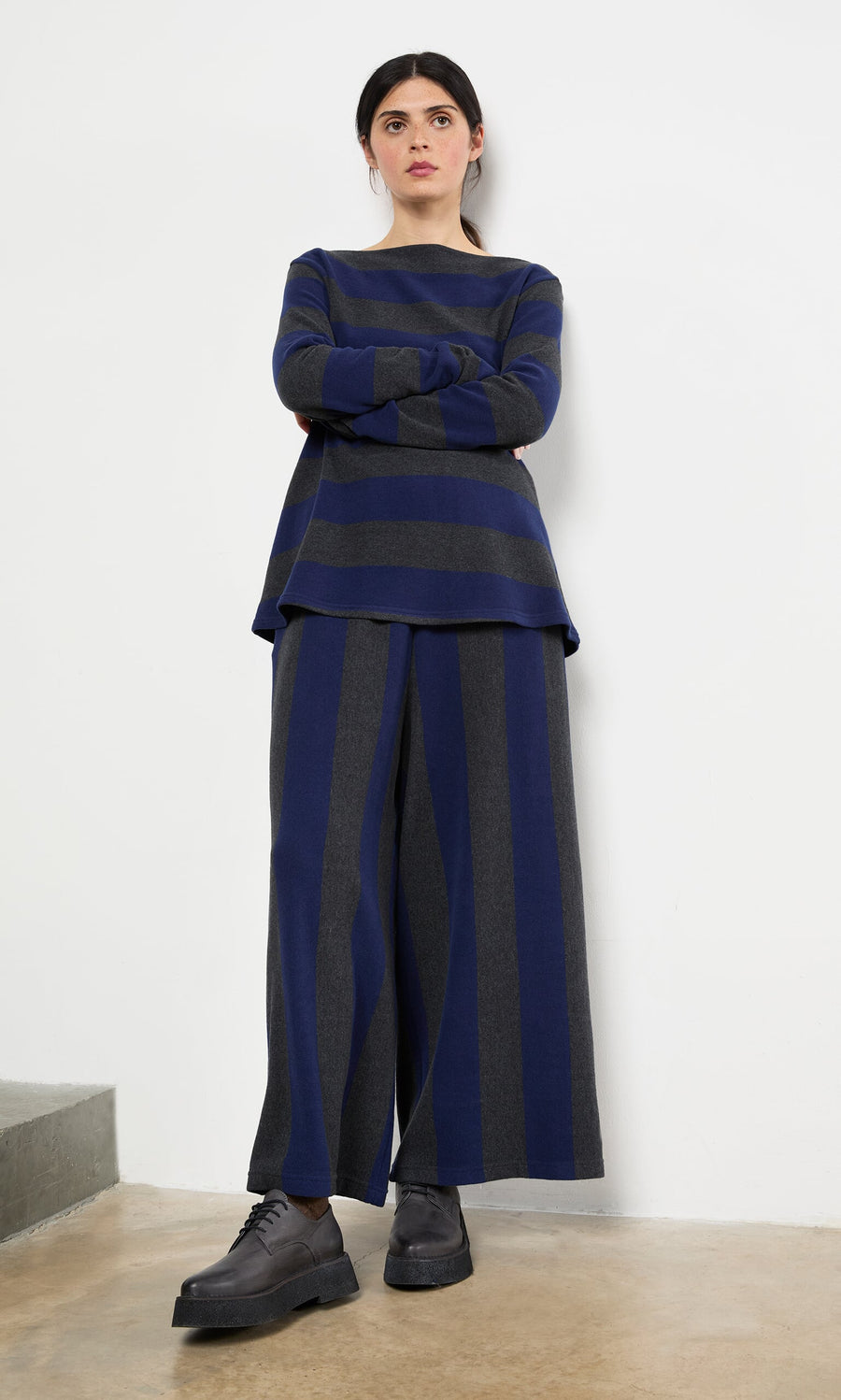 Plümo- Woman standing against a white wall wearing Clara Stripe Pants and a loose striped top with black shoes.