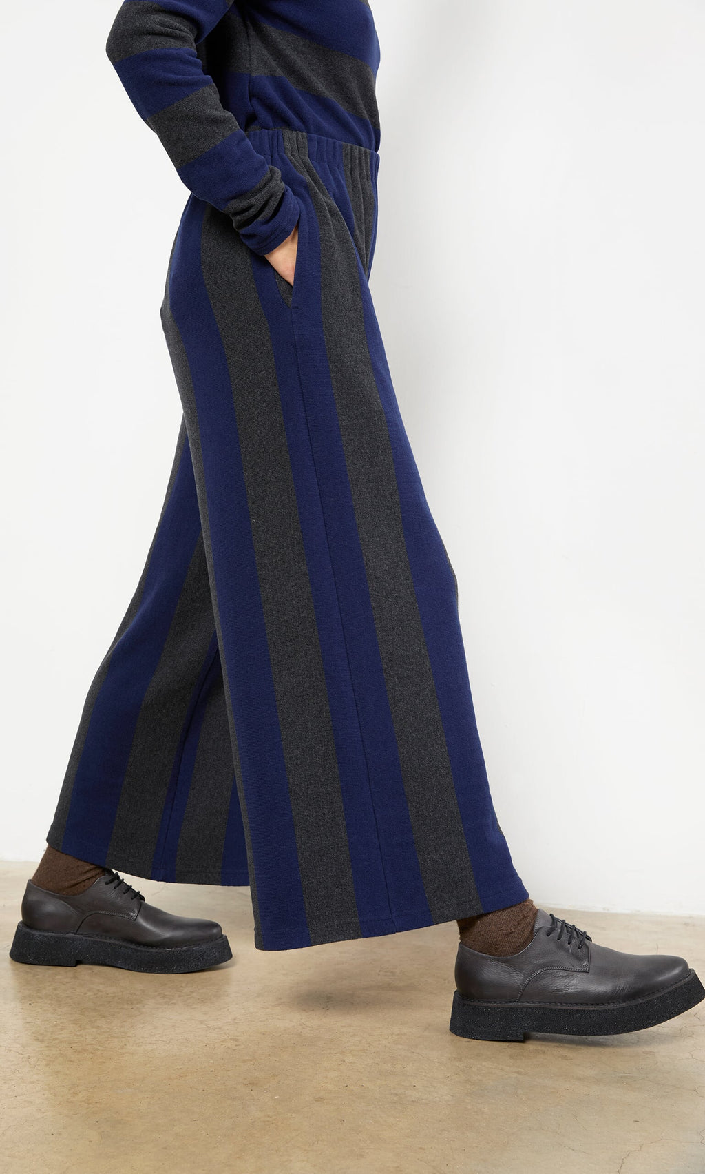 Plümo- Person wearing Clara Stripe Pants with brown socks and black shoes, standing on a concrete floor.