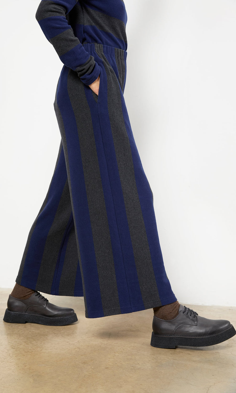 Plümo- Person wearing Clara Stripe Pants with brown socks and black shoes, standing on a concrete floor.
