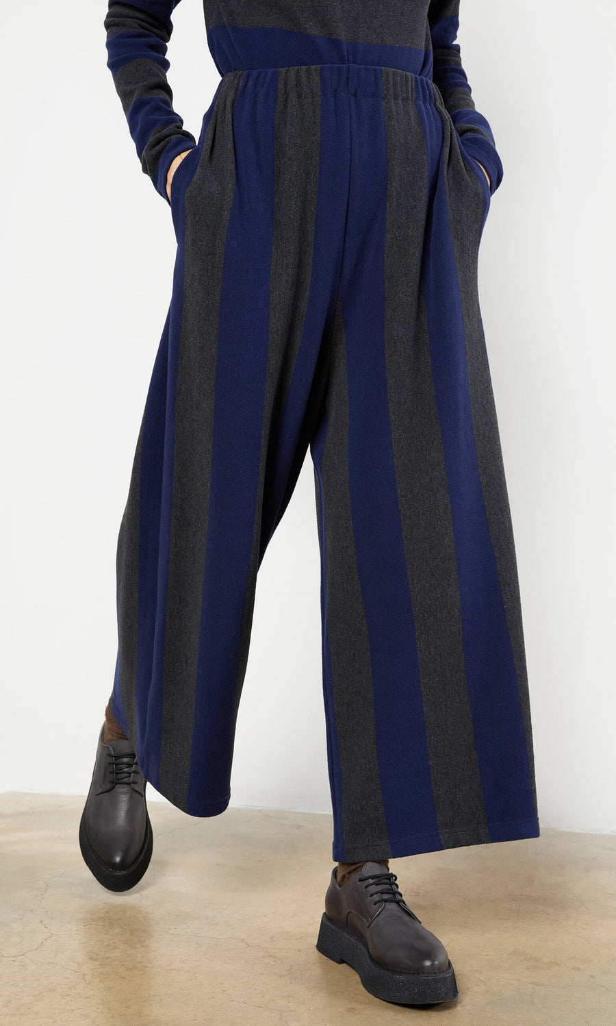 Plümo- Person wearing Clara Stripe Pants—wide-leg, cropped, vertical stripes—with dark shoes and a matching long-sleeve top indoors.