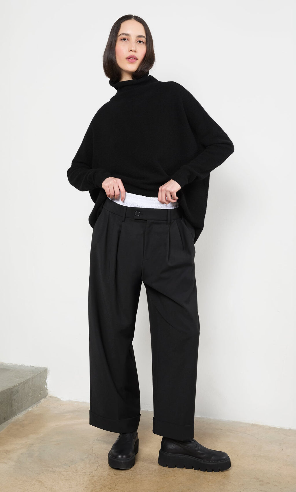 Plümo- Woman in Garconne Pants, a black turtleneck, and wide leg trousers stands indoors adjusting her waistband.