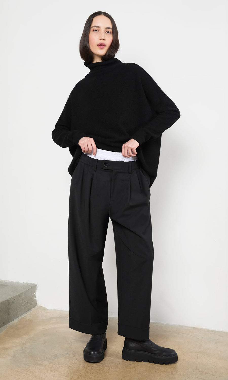 Plümo- Woman in Garconne Pants, a black turtleneck, and wide leg trousers stands indoors adjusting her waistband.