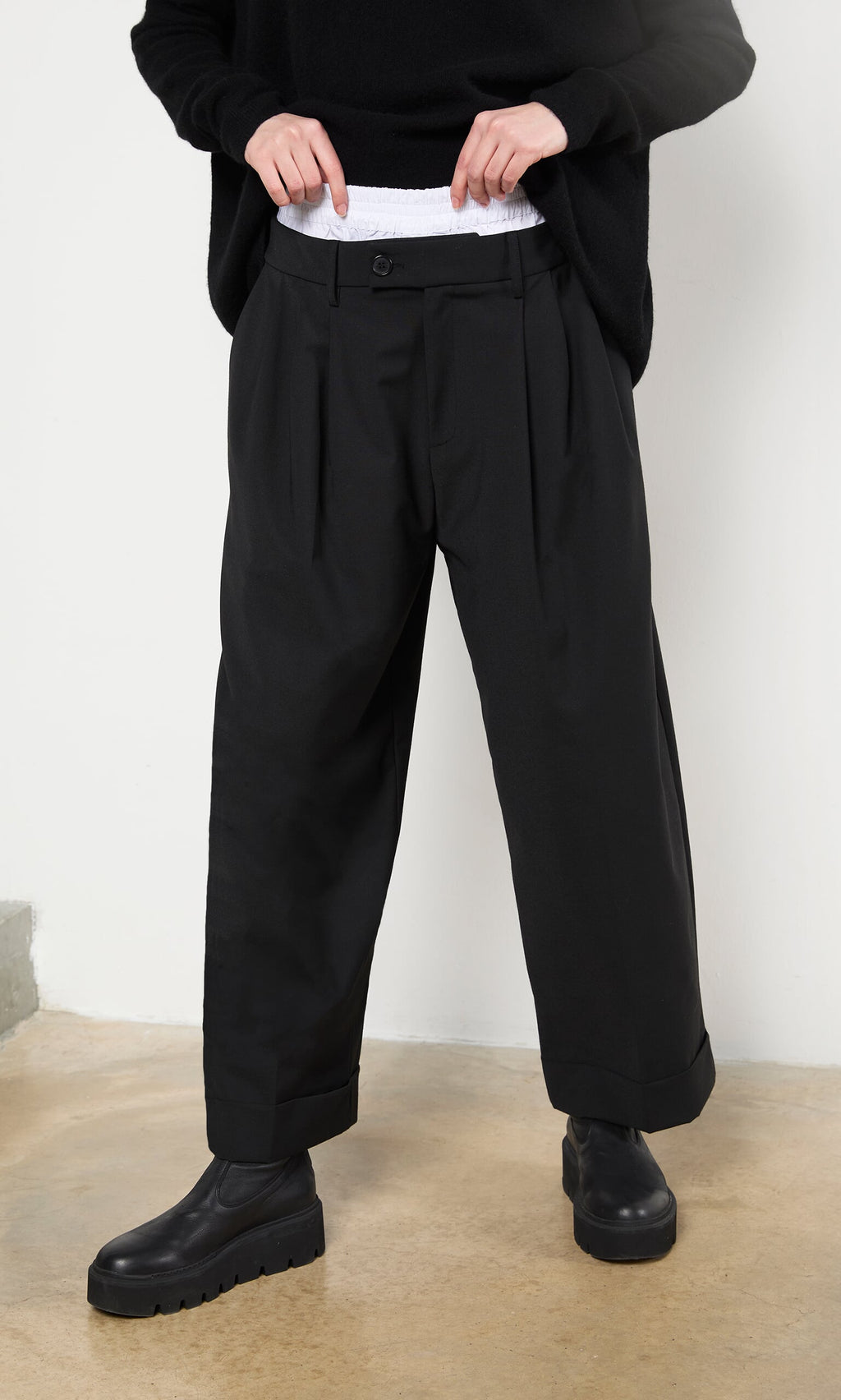 Plümo- A person wears Garconne Pants, a slouchy top, and black shoes, standing on a tan floor by a white wall.