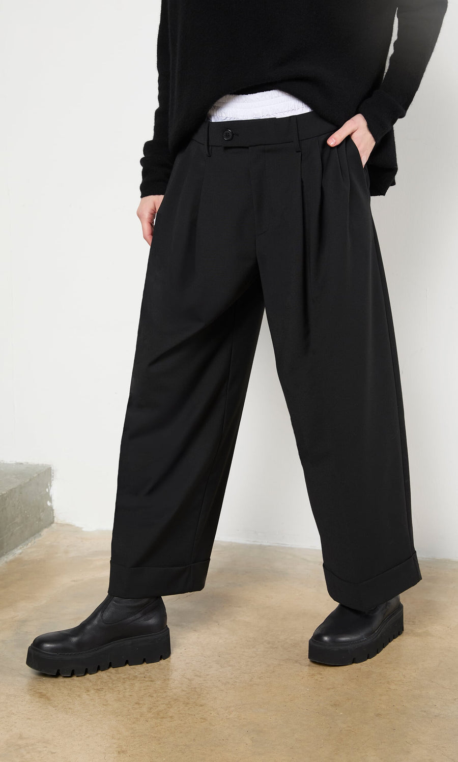 Plümo- Person wearing Garconne Pants, a black sweater, and platform shoes, standing on a concrete floor indoors.