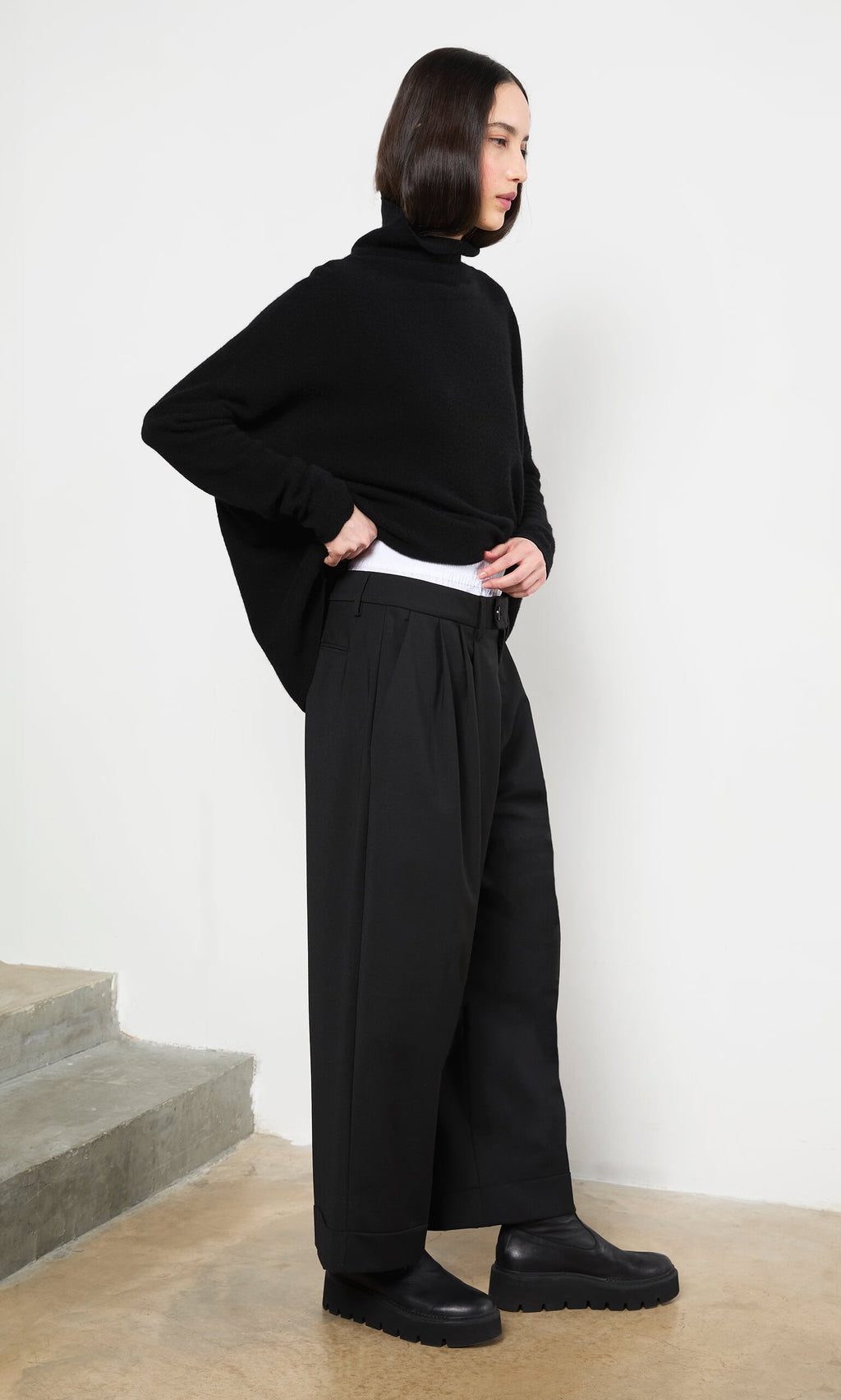 Plümo- Woman in Garconne Pants and a black turtleneck stands indoors by stairs, looking to the side.