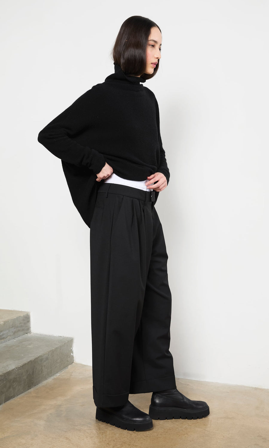 Plümo- Person standing indoors in an Onyx Rollneck sweater—a timeless classic—by concrete steps.