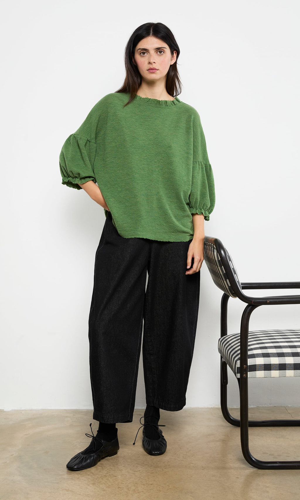 Plümo- Woman in a green Hopper Blouse and black wide-leg pants stands beside a black-and-white checkered chair.
