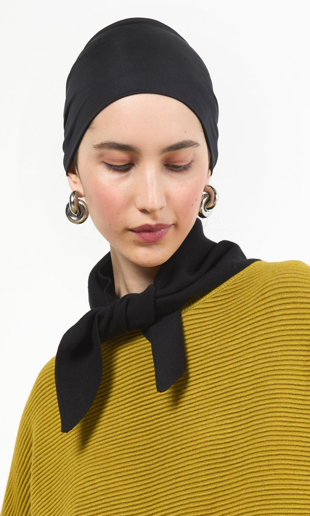 Plümo- Woman wearing a Black Wide Headband, bold earrings, and a mustard yellow textured top, looking down thoughtfully.
