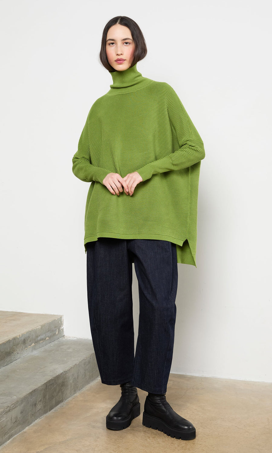 Plümo- Woman in the Jack Rollneck - Apple Green, wide-leg dark jeans, and black boots stands by concrete steps indoors.