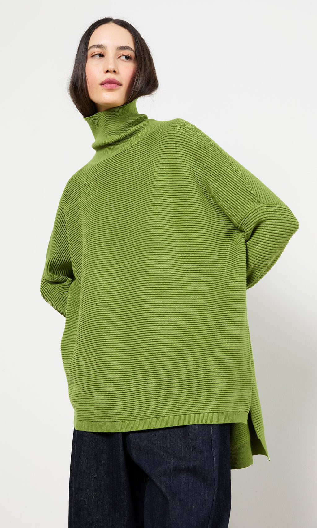 Plümo- Woman in the Jack Rollneck - Apple Green sweater and dark pants, standing against a plain white background.