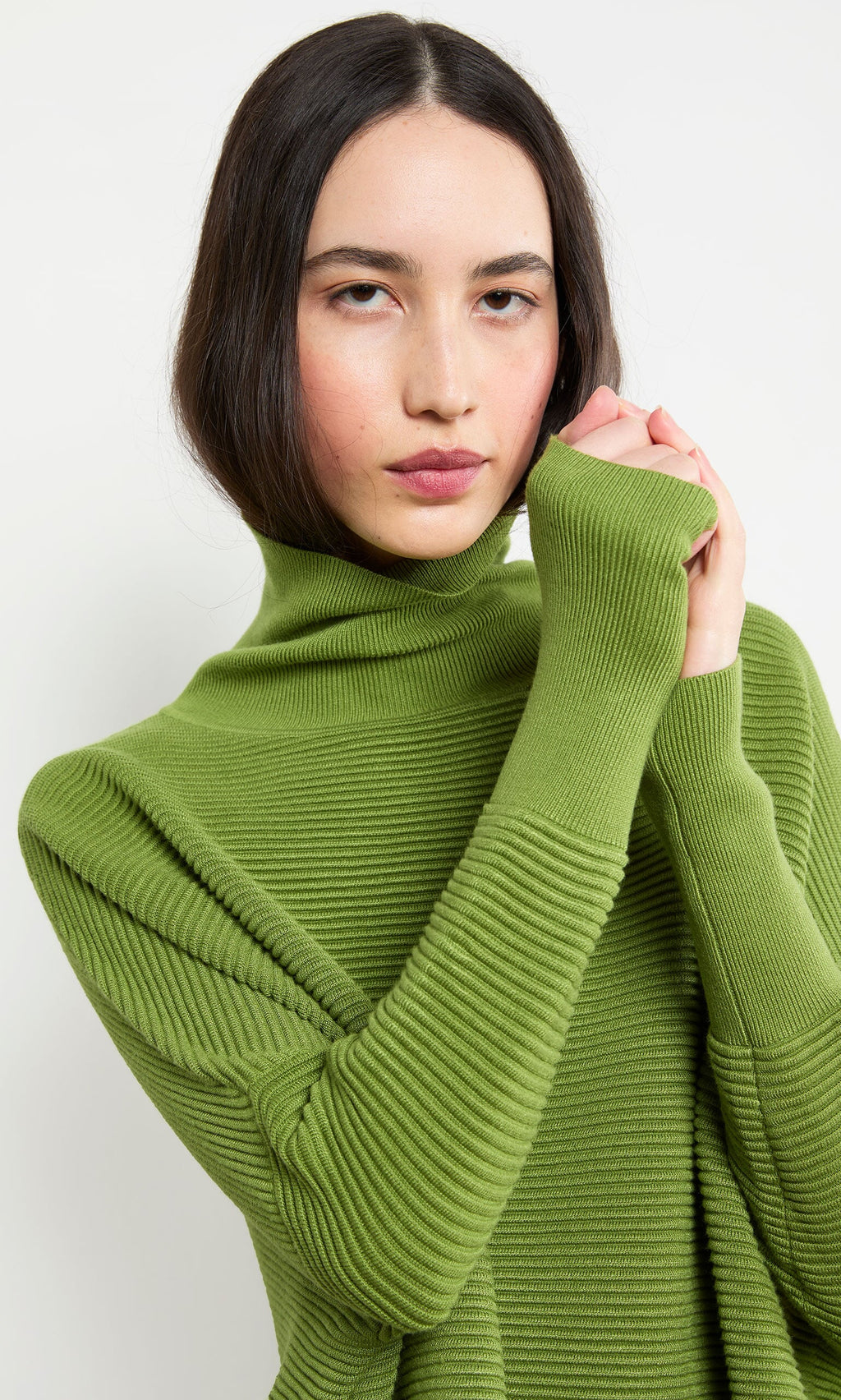 Plümo- Woman wears the Jack Rollneck in Apple Green, standing against a white background with hands near her face.