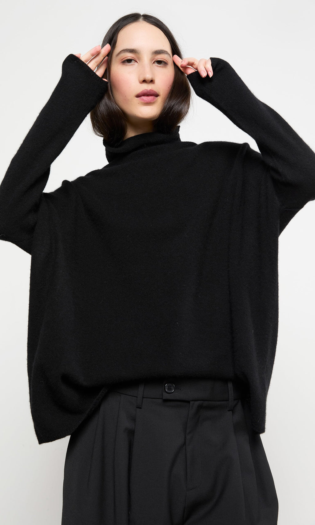 Plümo- Woman wearing the Onyx Rollneck and pants, posing with hands near her head against a white background.