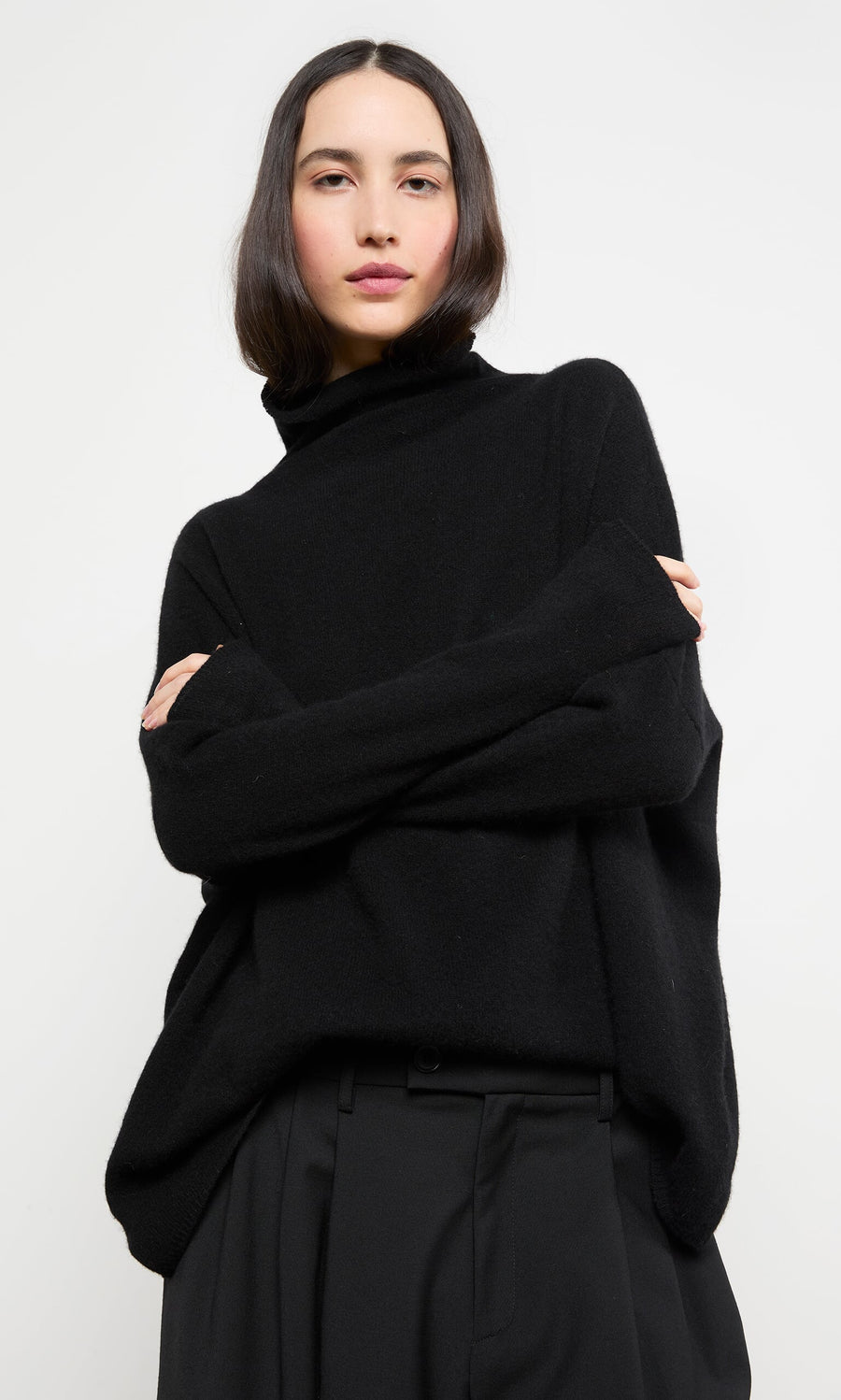 Plümo- Woman with straight dark hair wears the Onyx Rollneck sweater and black pants, standing against a white background.