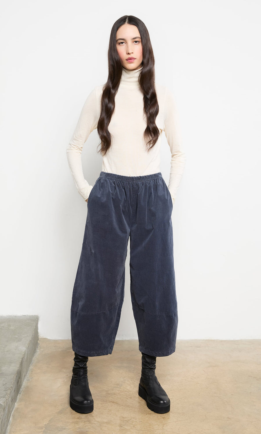 Plümo- Woman with long dark hair wears the Gentle Turtleneck, wide navy pants, and black boots against a plain background.