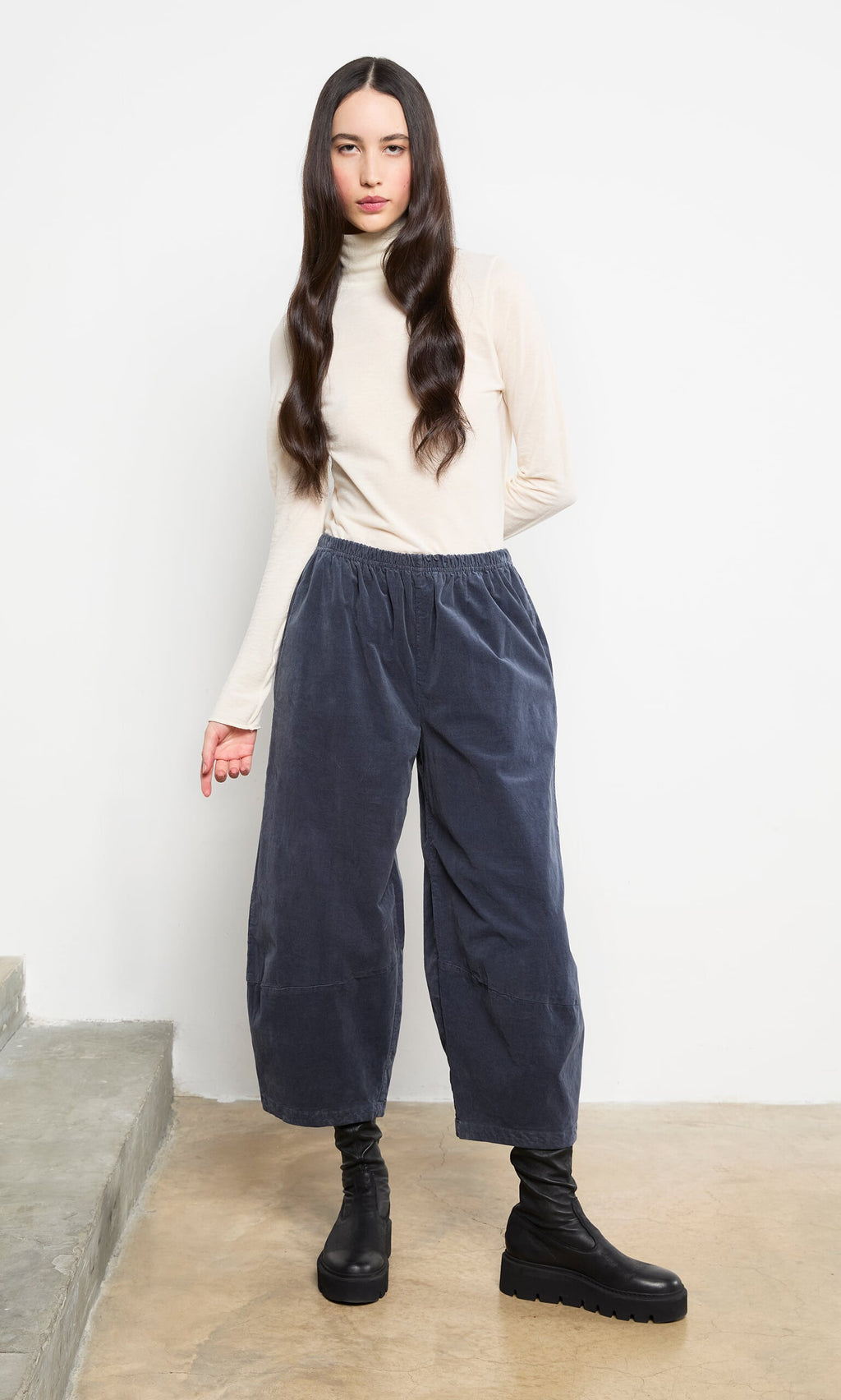 Plümo- A woman in a Gentle Turtleneck and gray pants stands indoors, wearing black boots with long, wavy hair.