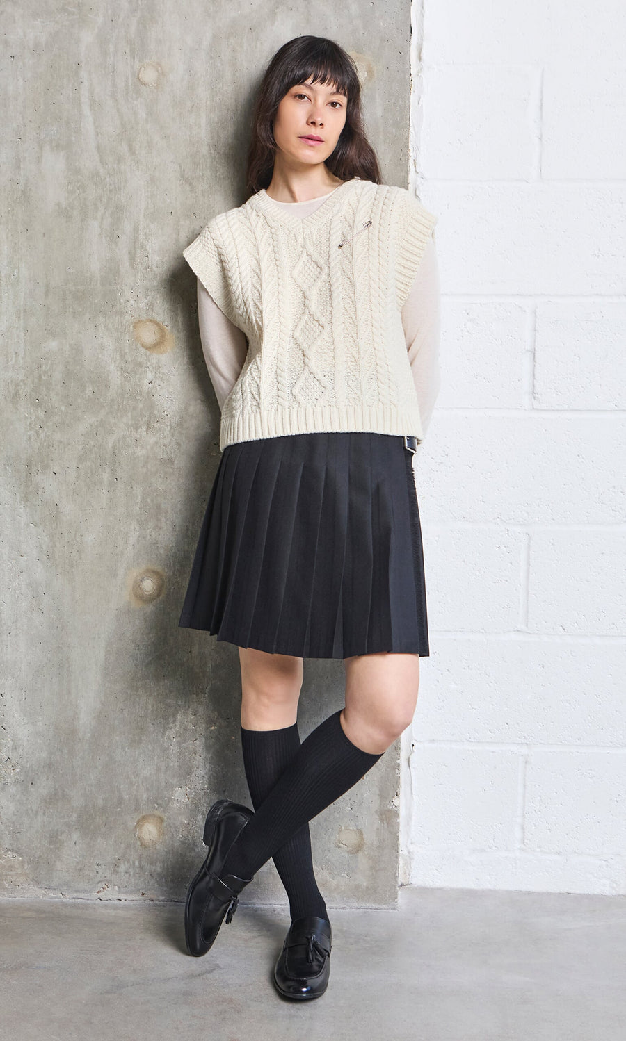 Plümo- Woman in a cream cable knit sweater vest, short black kilt, black knee socks, and loafers standing by a wall.