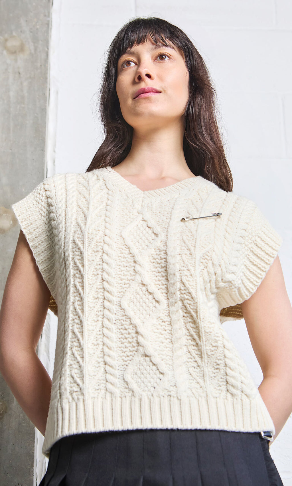 Plümo A woman styles a cream cable knit sleeveless sweater with a Kilt Pin, standing by a concrete and white wall.