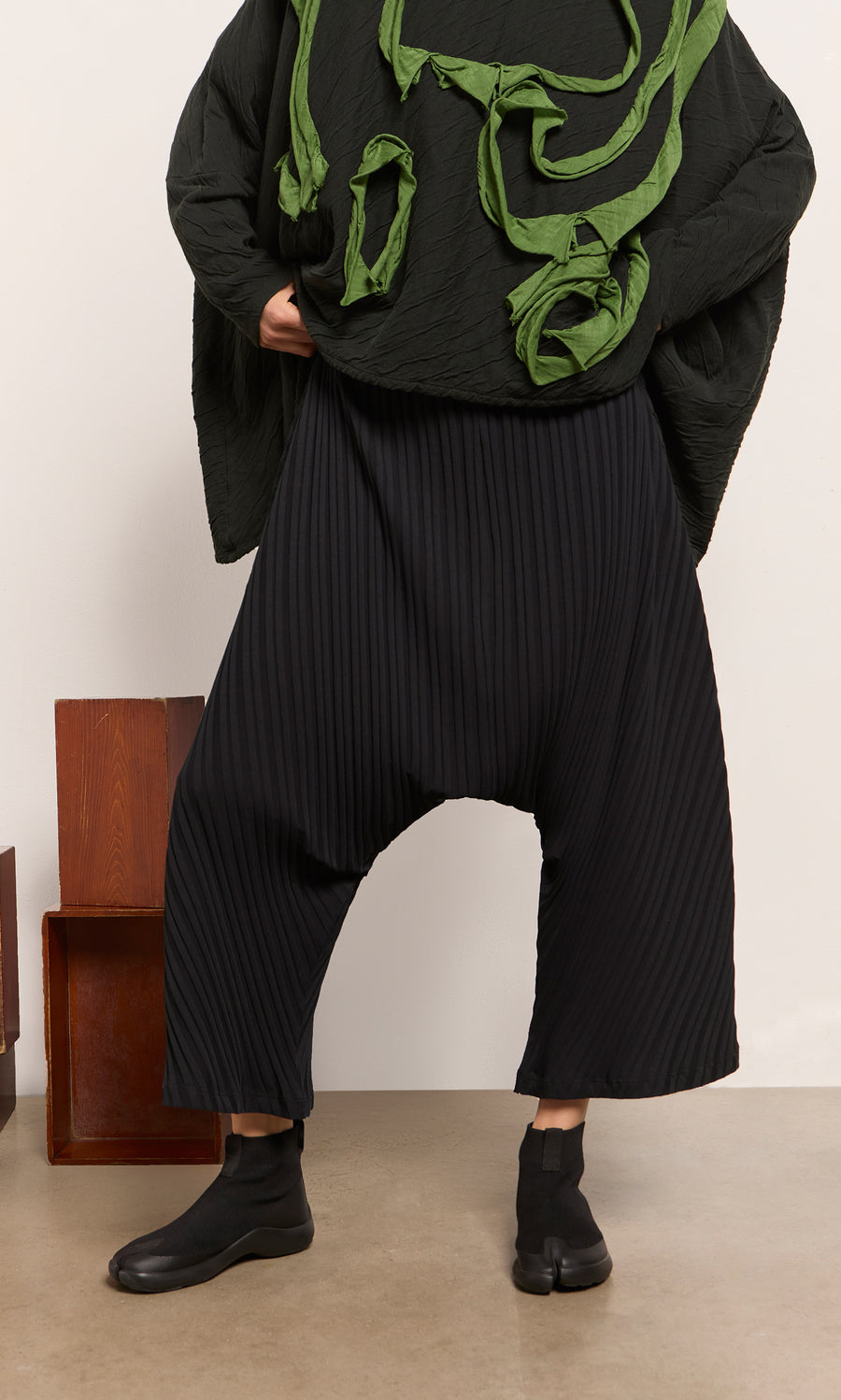 Plümo  Person in black Acker Pants, textured Danish design top, and black shoes stands indoors.