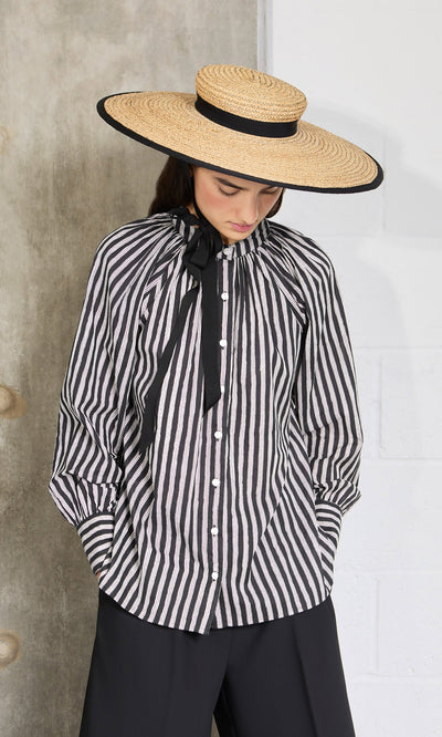 Plümo- Woman in a wide-brimmed straw hat and puffed sleeves wears the Ailsa Blouse - Charcoal Stripe, looking down.