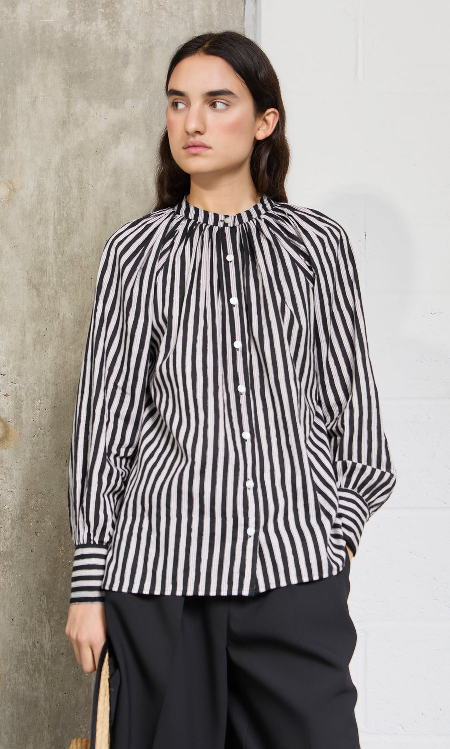 Plümo- A woman in the Ailsa Blouse - Charcoal Stripe with black pants stands by a concrete and white wall, looking to the side.