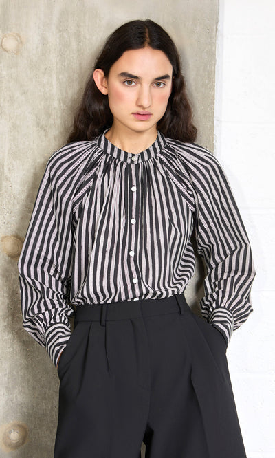 Plümo- Woman standing against a concrete wall in the Ailsa Blouse - Charcoal Stripe and high-waisted black trousers.