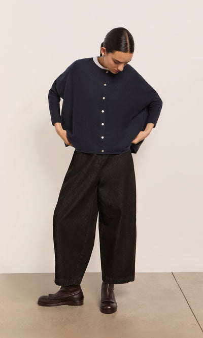 Plümo  Woman in the Alma Cardigan - Dark Navy, wide black pants, and brown boots stands looking down with hands in her pockets.
