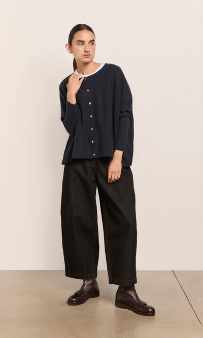 Plümo  Person wears the Alma Cardigan - Dark Navy over a white shirt, black pants, and brown boots against a plain wall.