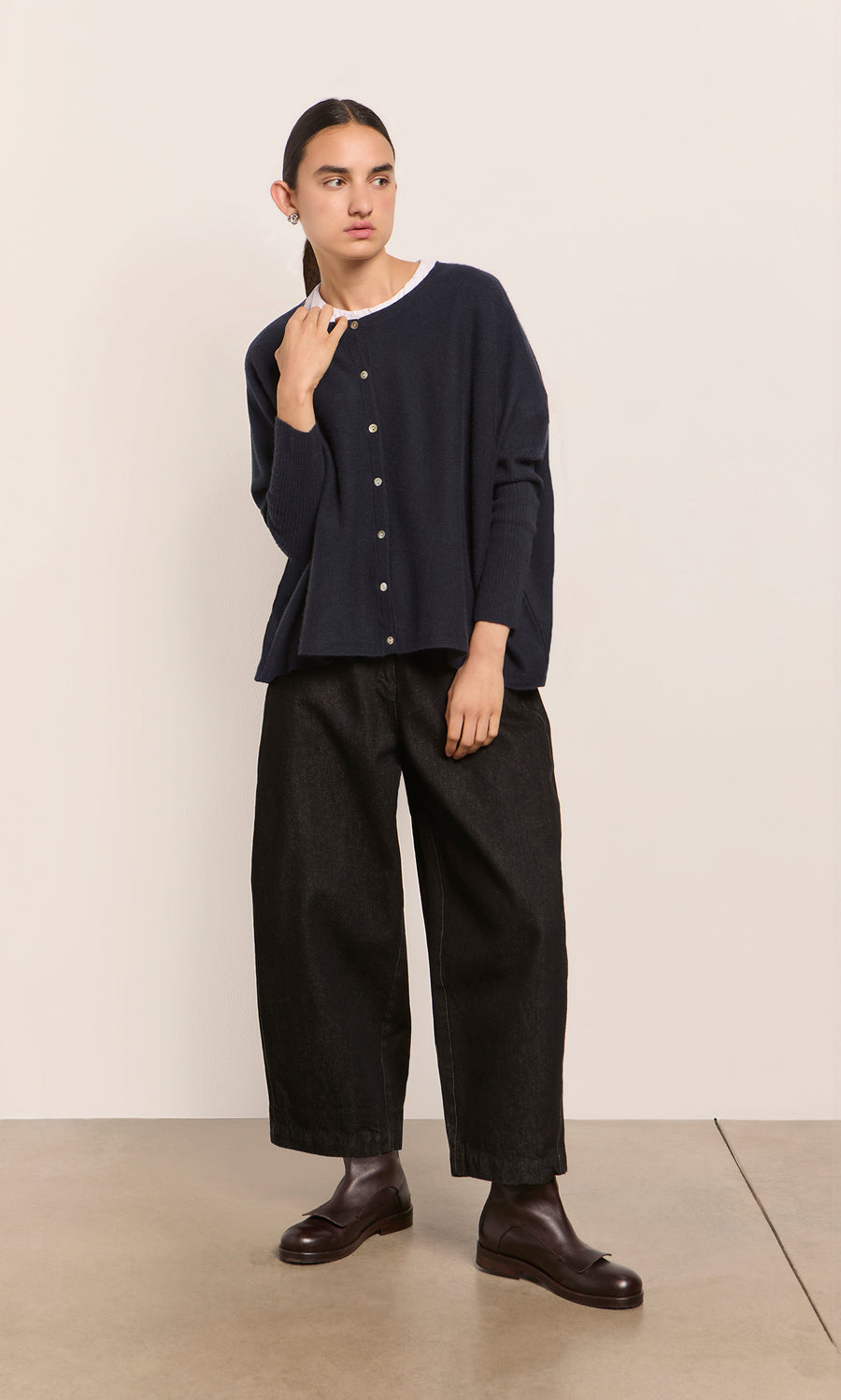 Plümo  Person wears the Alma Cardigan - Dark Navy over a white shirt, black pants, and brown boots against a plain wall.