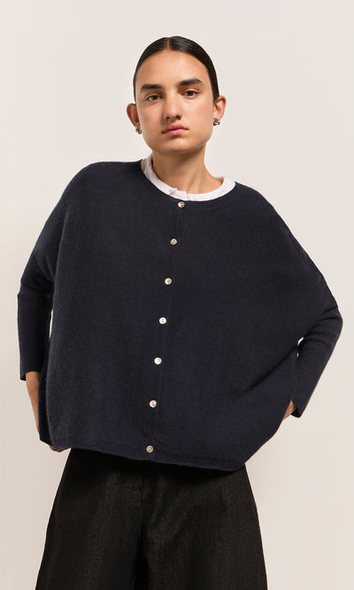 Plümo  Model in Alma Cardigan - Dark Navy over a white shirt, standing against a plain, light-colored background.